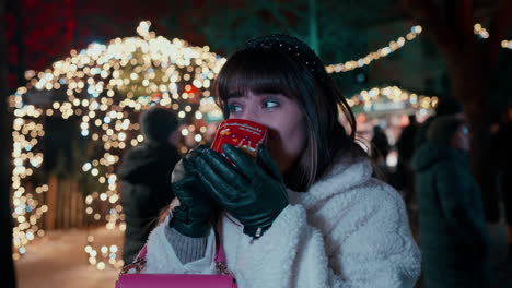 beautiful woman with dark hair saying cheers with her hand and drinking punch from red heart cup at vienna christmas market , surrounded by christmas lights in the background