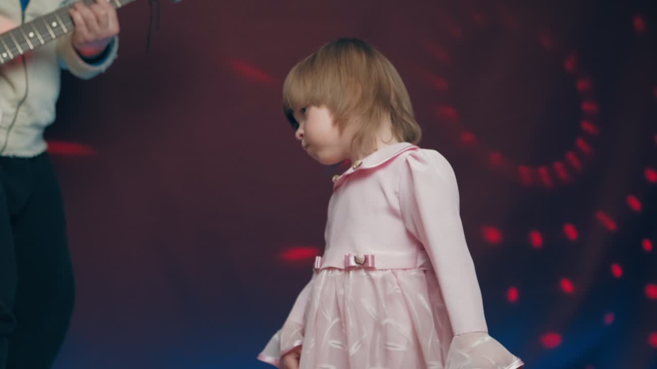 una niña con un vestido vintage baila en el escenario, su padre toca la guitarra acústica