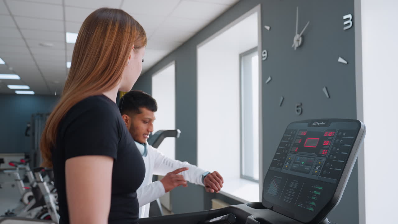 woman focusing on treadmill session with male trainer nearby checking time as treadmill operates under large wall clock in bright gym interior with modern decor and visible digital display panel