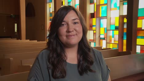 a pretty young ethnic female smiles at the camera inside a church sanctuary while sitting in a pew