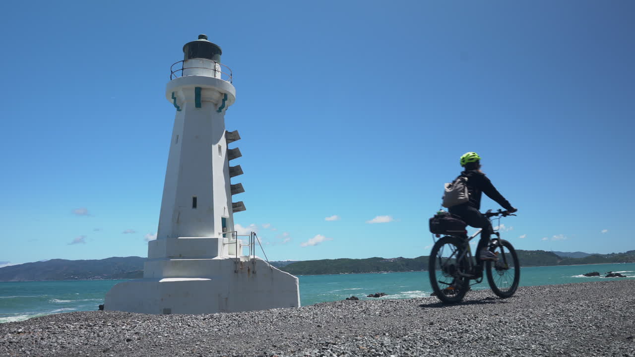 A Female cyclist rides past Pencarrow Lighthouse on a sunny day on an E-bike