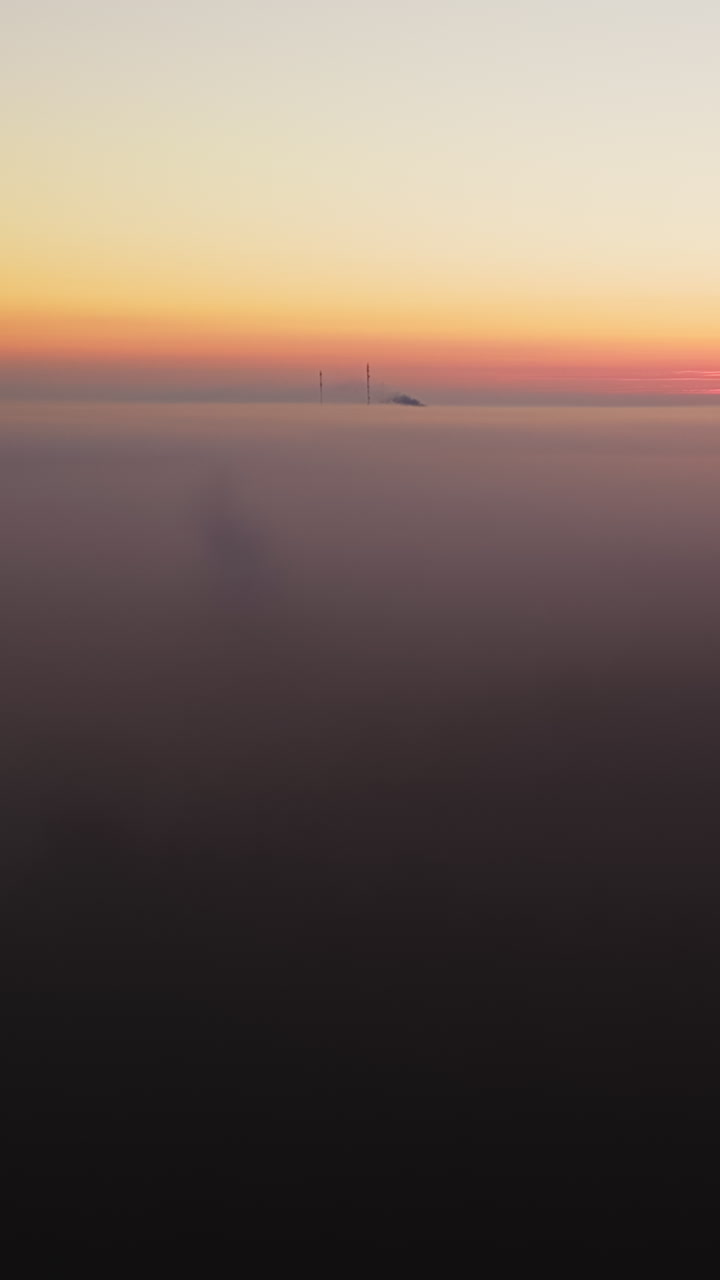 Vertical drone shot rising over dense fog, toward radio towers, vibrant sunrise