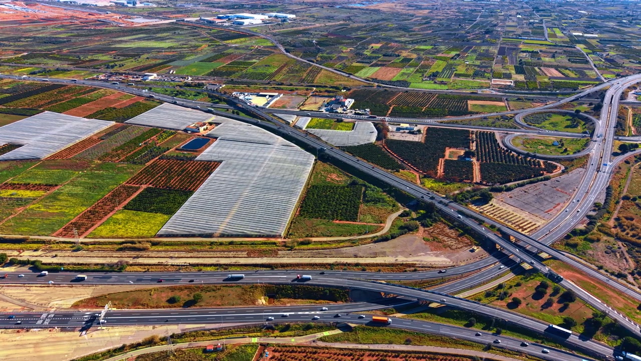 Luchtfoto van een snelwegknooppunt door landbouwvelden en kassen. Breed landschap met een meerbaansweg die langs kassen en sinaasappelboomgaarden loopt in een landelijk gebied onder een felle zon.