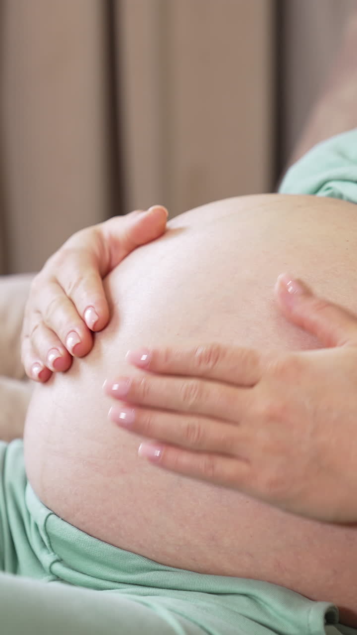 Caring future mother stroking her big tummy while resting on the sofa. Woman caressing her child in the big belly. Two little baby socks are on the armrest at he backdrop. Vertical video