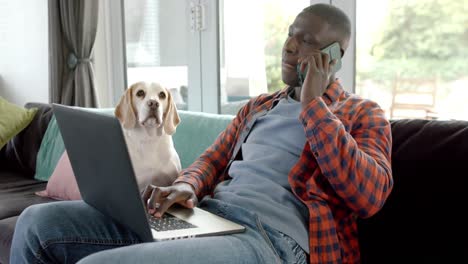 African-american-man-talking-on-smartphone-and-using-laptop-at-home,-with-his-pet-dog,-slow-motion