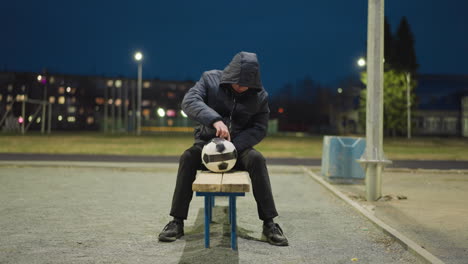 a man is seated on a bench at night, thoughtfully rotating a soccer ball between his legs, with blurred lights and goalposts in the background