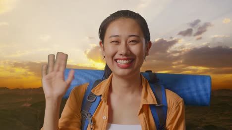 close up de una excursionista asiática con mochila de montañismo agitando la mano y diciendo adiós mientras está de pie en la cima de la montaña durante la hora de la puesta del sol