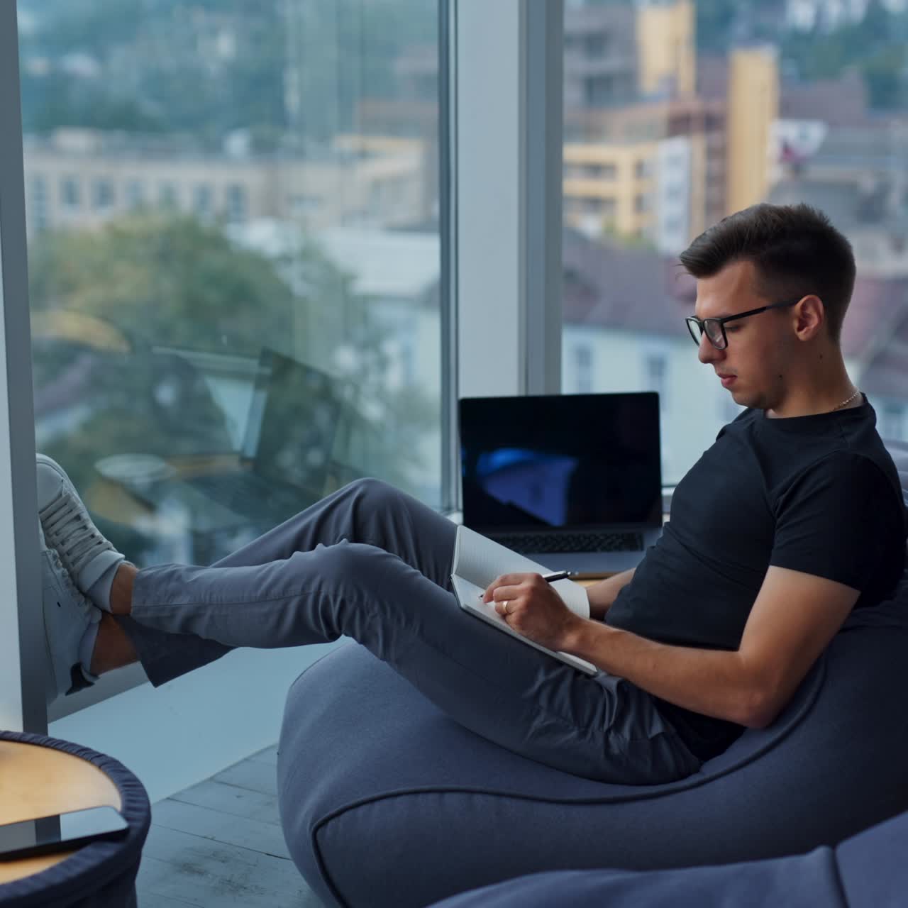 Coming closer to a young man sitting relaxed near the window. Man wearing glasses writing something in paper book. Blurred backdrop