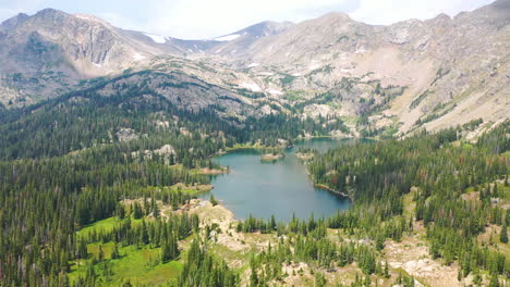 Imágenes-Aéreas-De-Drones-Del-Hermoso-Lago-De-Montaña-Rocosa-Con-Agua-Azul-Clara-Rodeada-De-Picos-Montañosos-Y-Un-Espeso-Bosque-De-Pinos-En-Nederland-Colorado