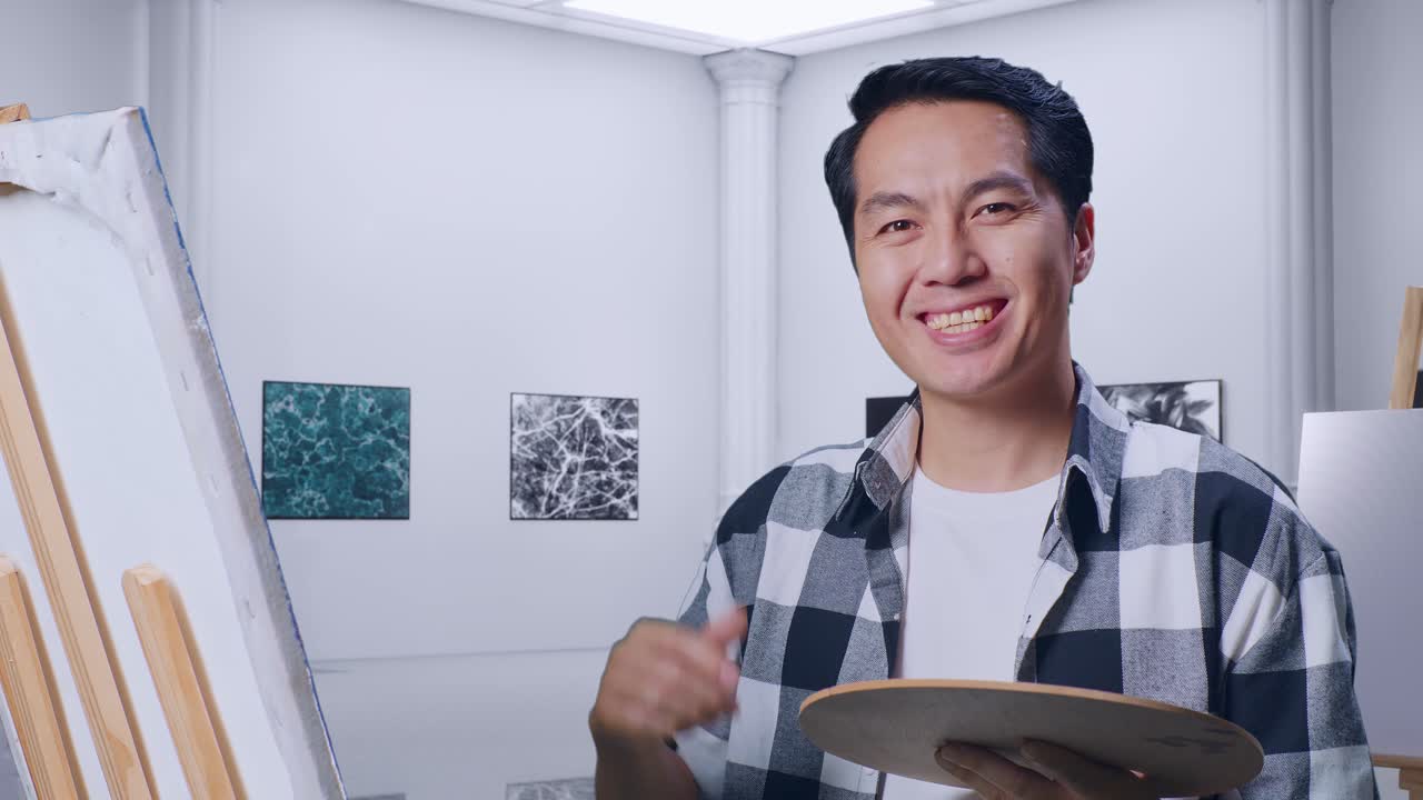 アジアの男性アーティストが画廊で油絵とブラシでキャンバスに絵を描く際に親指を上げてジェスチャーをして笑顔を浮かべています