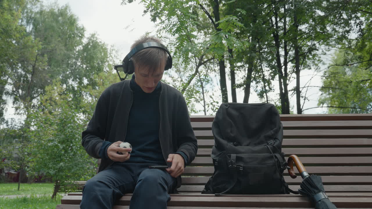 pria di bangku, headphone, ransel, payung, inspeksi gadget kecil, latar belakang taman mendung dengan pepohonan, gerakan lambat dan hati-hati, saku dan tangan menyesuaikan perangkat, suasana hati yang introspektif dan halus