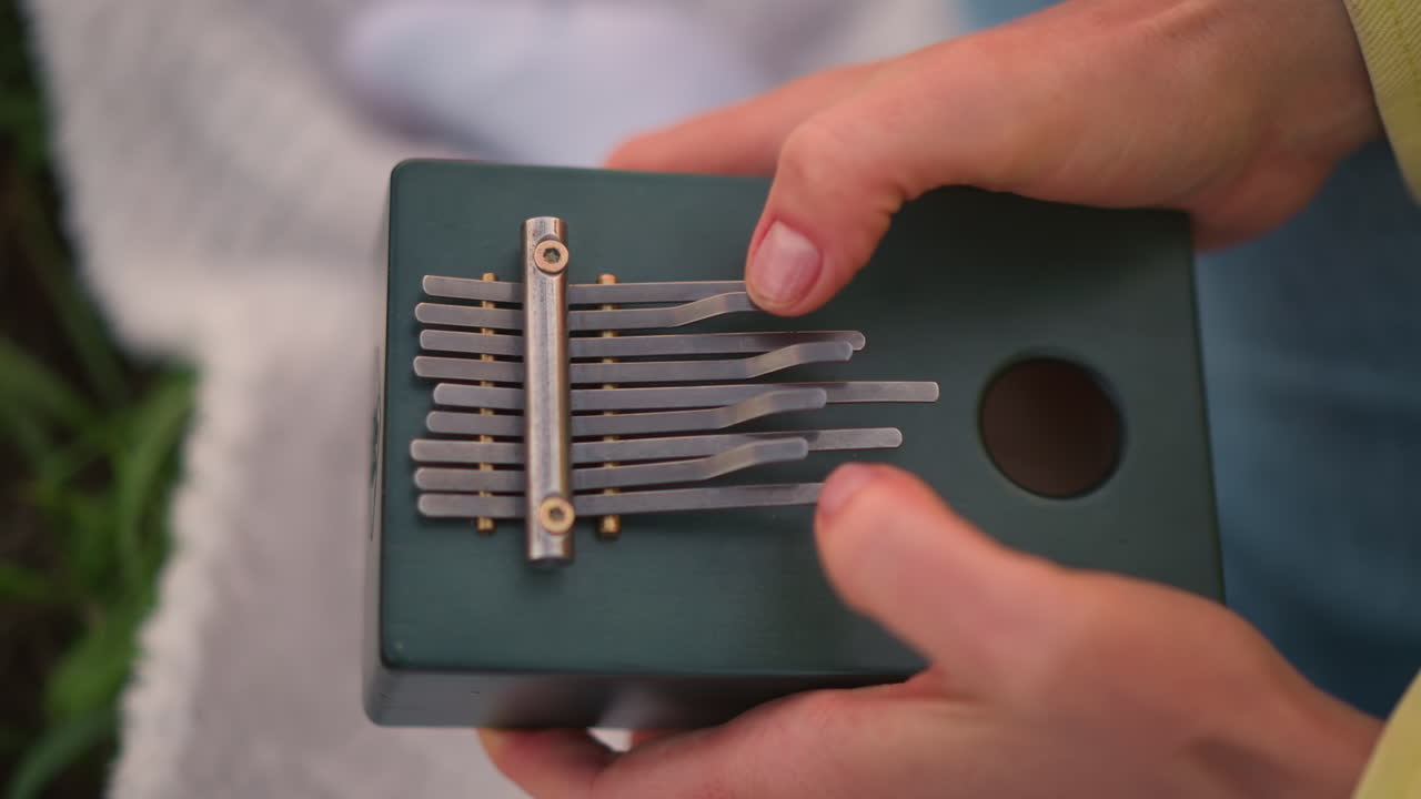 담요 위에서 흰 손으로 초록색 칼림바를 조율하는 모습이에요. 원형 사운드 홀이 있는 상자 악기의 클로즈업 샷이며, 섬세한 엄지 손가락 연주와 정확한 조율, 연습과 선생님을 위한 교육적인 분위기가 느껴져요.