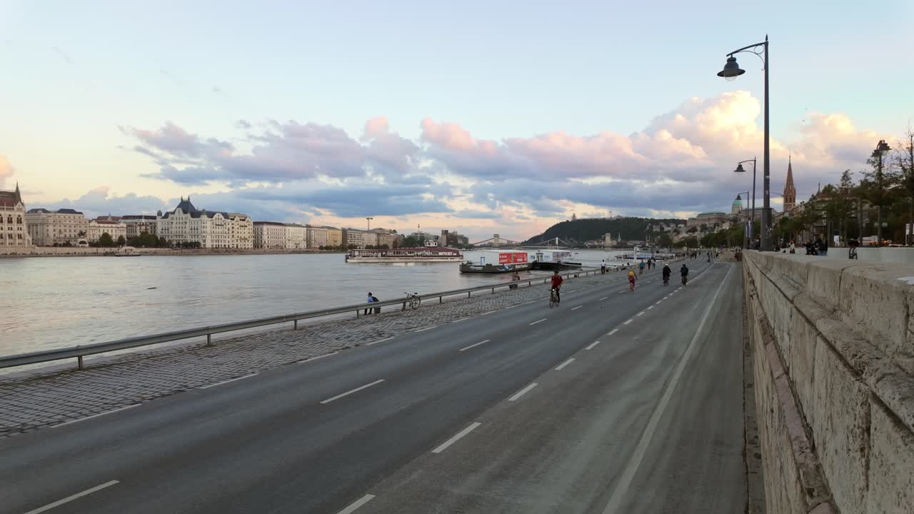 People walk, run and cycle on the lower Buda quay, which is closed due to the flooding of the River Danube and Budapest's landmarks in the background at twilight.