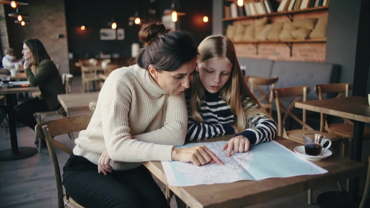 Assistente adulto con maglione che indica una mappa aperta e guida una ragazza in un accogliente caffè, pianificando il percorso accanto a una tazza.
