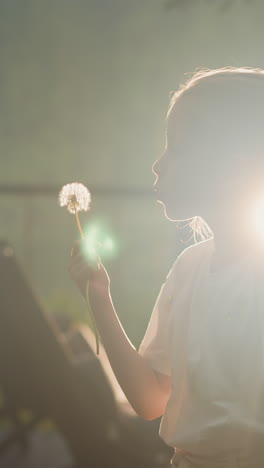 child blows seeds from dry dandelion. girl enjoys flowers standing against bright sun in yard of country house side view slow motion. vacation in natural environment