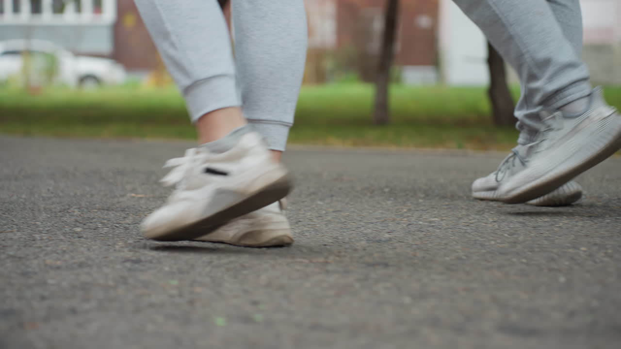 Side leg view of athletes walking side by side wearing sneakers and joggers on paved street, with slightly blurred background showing parked car, greenery, and building during outdoor stroll