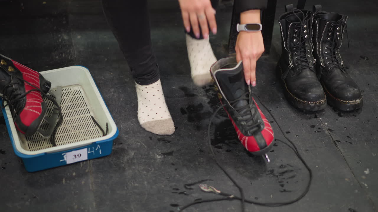 Female wearing polka dot socks holding pair of black boots on wet locker room floor with red and black ice skates resting inside blue tray after changing footwear during winter preparation indoors