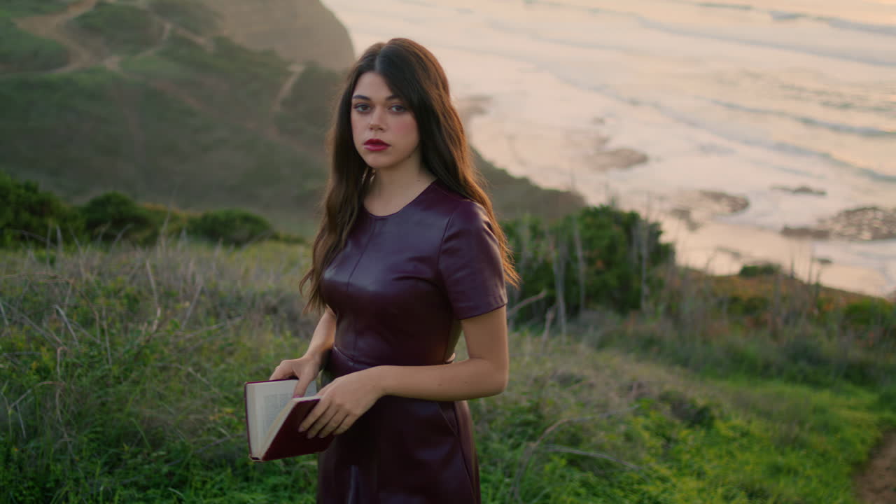 편안한 소녀가 바다 근처의 경사에서 책을 들고 다가오고 있습니다. 모델을 찾는 카메라