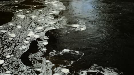 Lapso-De-Tiempo-De-Burbujas-Giratorias-De-Espuma-Blanca-Flotando-En-La-Superficie-Del-Agua-Oscura-En-Un-Río-Escocés-En-Un-Patrón-En-Constante-Cambio-Donde-La-Corriente-Más-Rápida-Se-Encuentra-Con-Una-Sección-De-Agua-Inmóvil