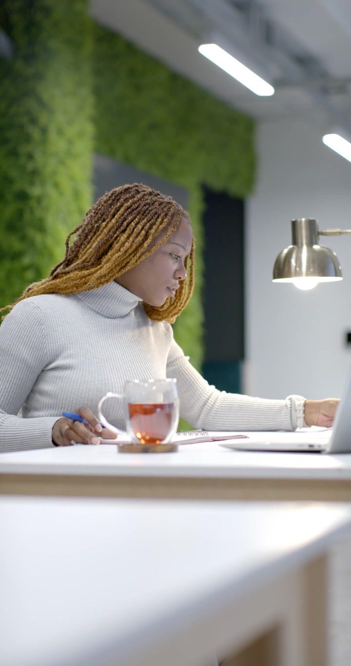 Vertical video: Glancing African American woman using laptop pulling page marking with pen at desk