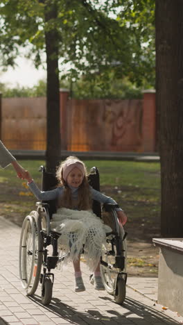 caring mother holds hand of daughter with spinal cord injury walking on stone pavement. little girl sits in wheelchair enjoying spring nature in park