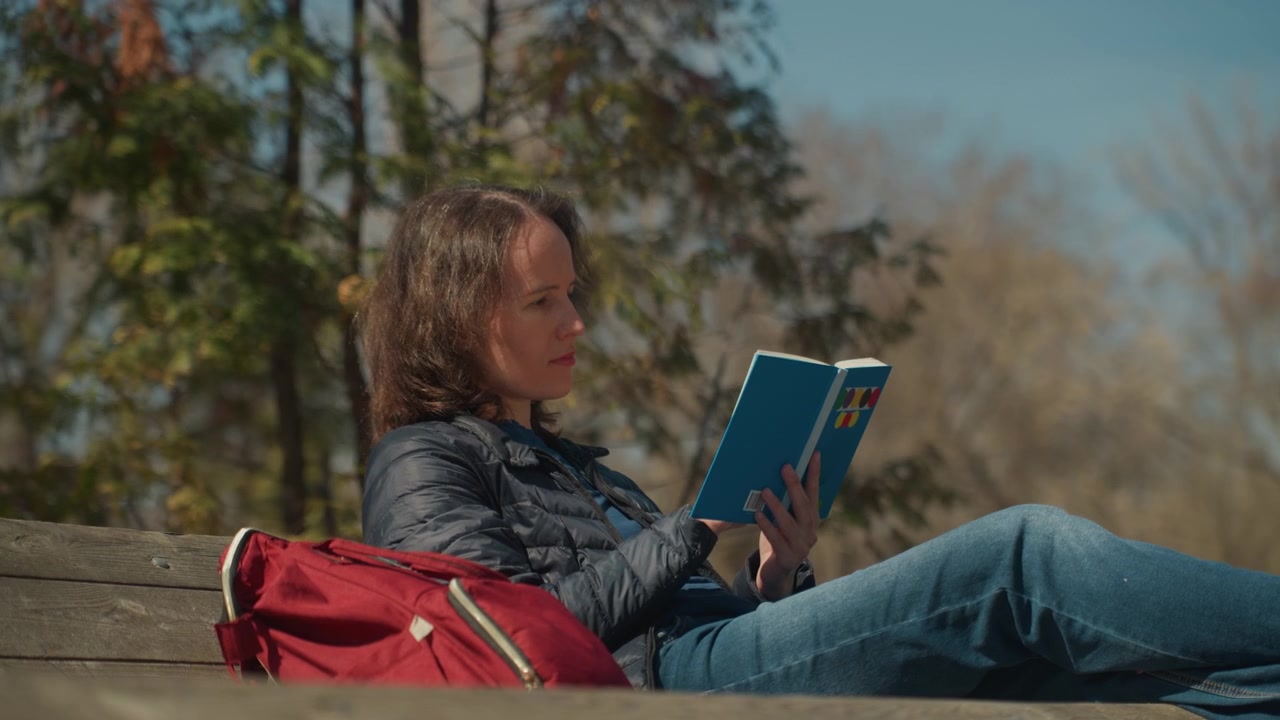 Close up of woman reading book on wooden bench with red backpack in outdoor park setting sunlight and trees visible closeup relaxed nature daytime leisure