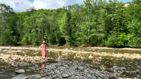 retro styled young woman in straw boater hat and long dress walking on river rocks near forest. romantic girl. natural scenic landscape. summer vacation. vintage aesthetic lifestyle. 4k video