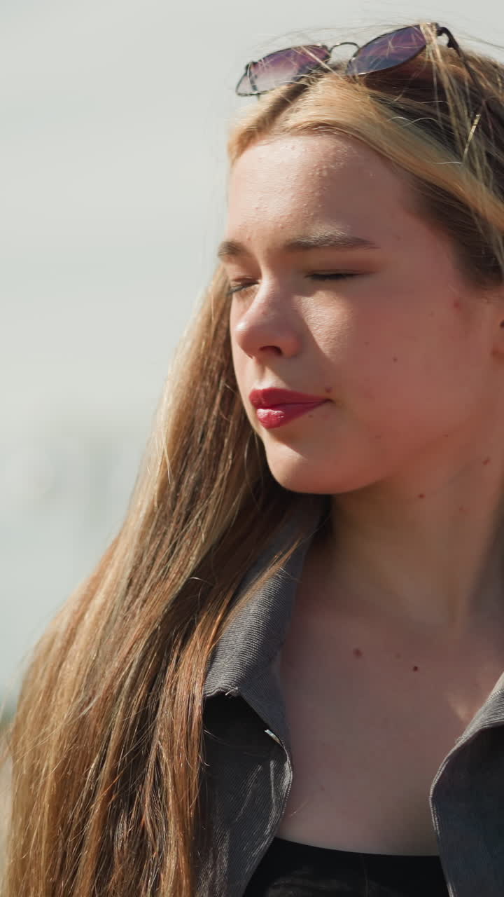 mujer con ropa gris ajusta su cabello a medida que sopla el viento, con los ojos ligeramente cerrados debido al brillante reflejo de la luz solar en su cara, el entorno al aire libre presenta un fondo borroso y postes de luz
