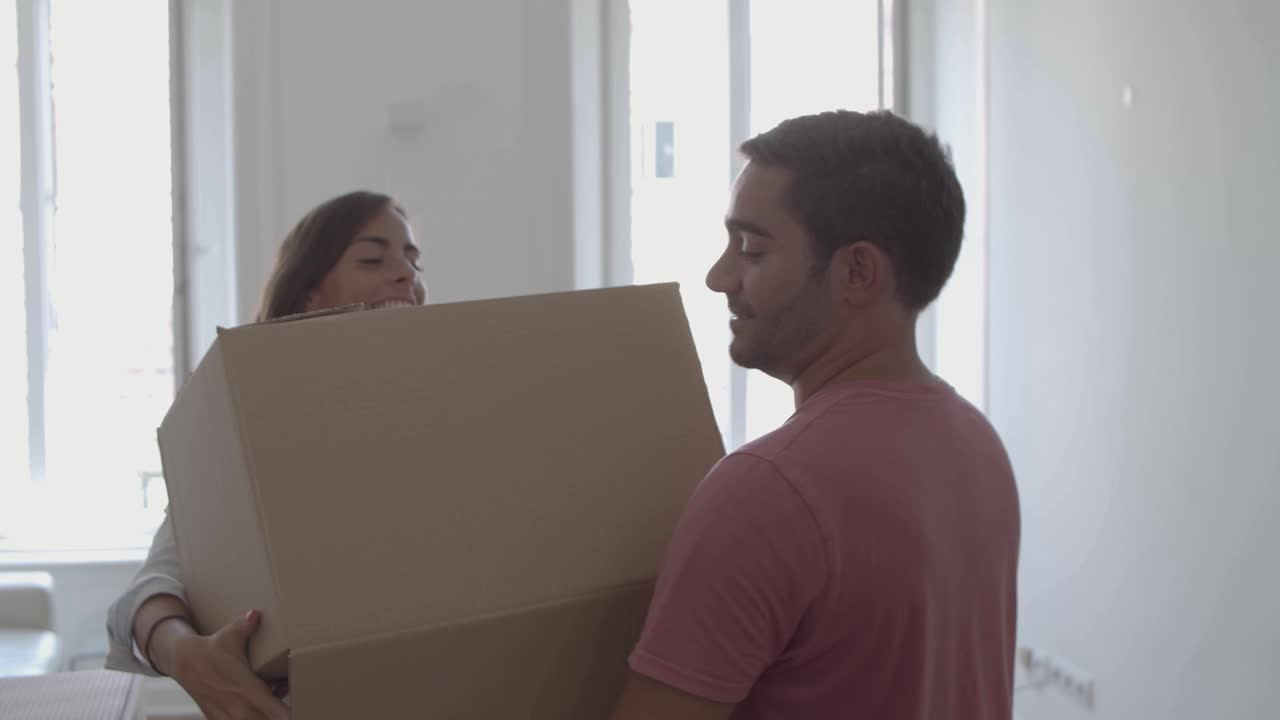 Happy man coming into room, carrying box with different stuff, kissing and hugging young wife