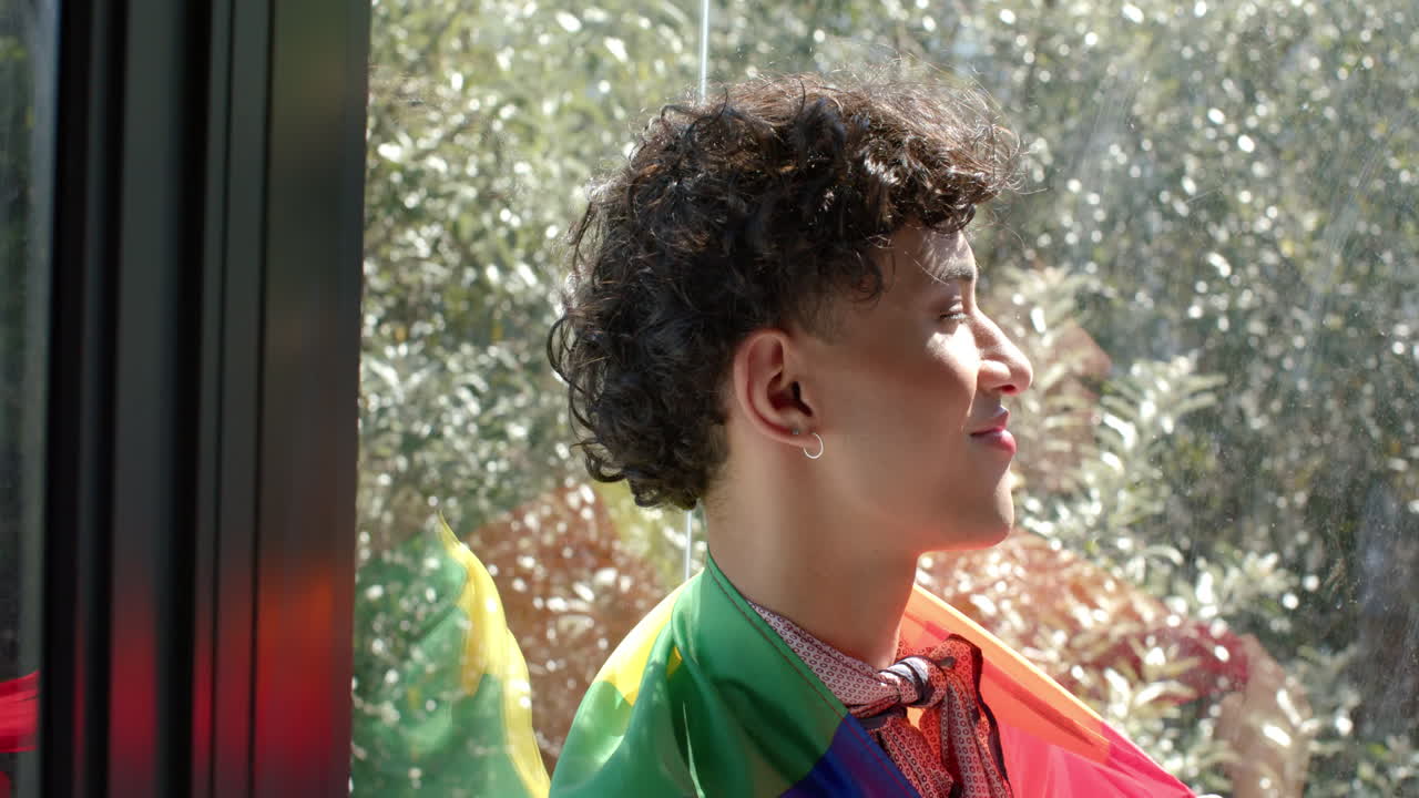 Smiling young person wrapped in rainbow flag, enjoying sunlight by window