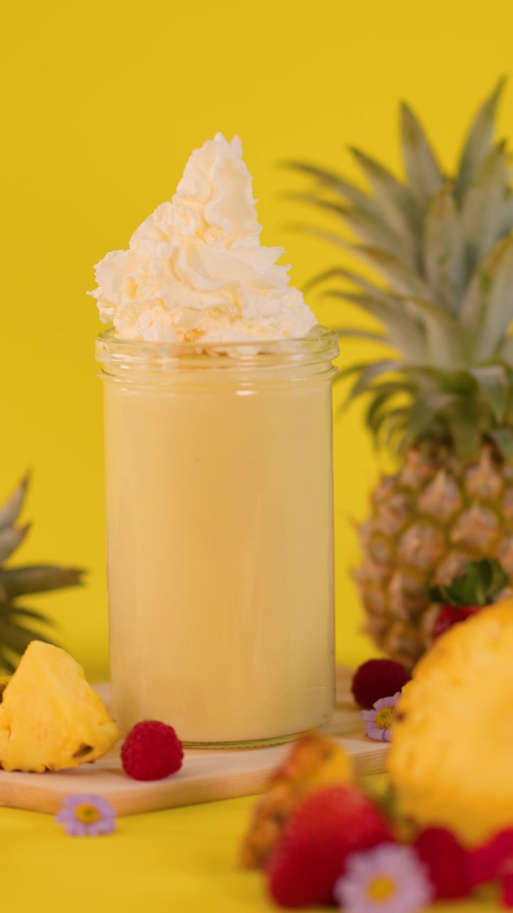 Una mano aggiunge panna montata a un frullato all'ananas con frutta fresca su uno sfondo giallo vivace.