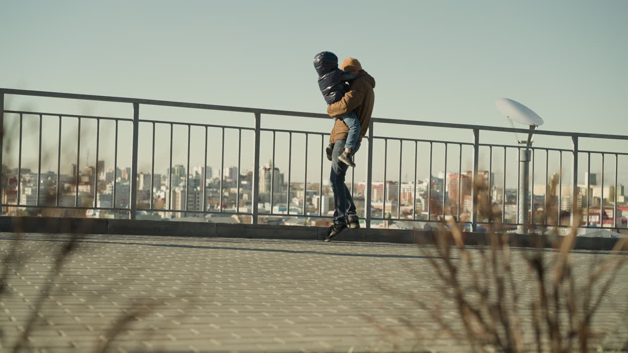 A father tenderly holds his son close to an iron railing, stepping down and then back up, both observing the cityscape below, they are both dressed jacket and beanie