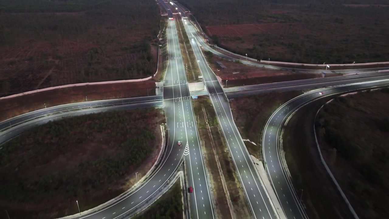 Wide cinematic aerial of an expansive expressway network showcasing curved access ramps, bright roadway illumination, and multilane transport corridors stretching into the night horizon.