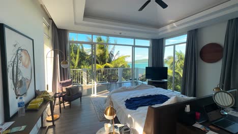 daytime interior of luxurious, slightly messy bedroom with king-sized bed and sunlit balcony overlooking palm trees and ocean