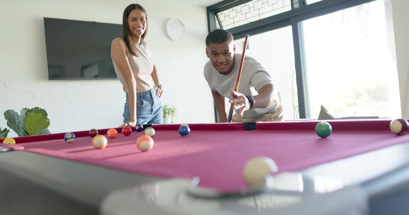 African American couple playing pool in living room, woman encouraging man aiming with cue stick