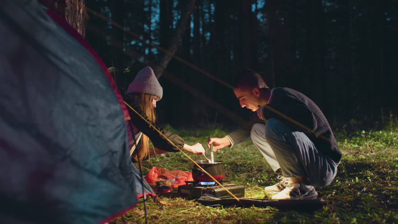 Couple prepares meal outside tent during night camping as man stirs pot and woman adds canned food, illuminated by phone light, surrounded by trees in forest with portable stove on grassy ground