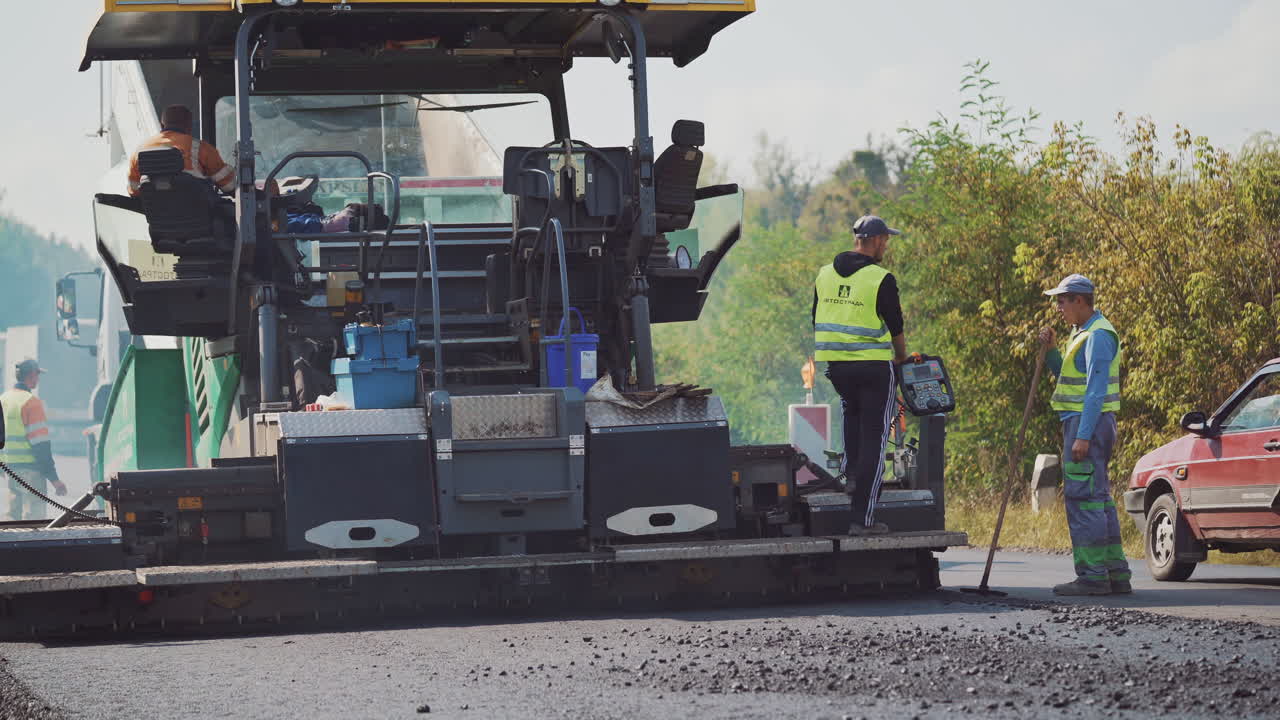 Asphalt paver machine and workers during the road construction. Male workers straighten black bitumen with special instruments on the background of traffic.