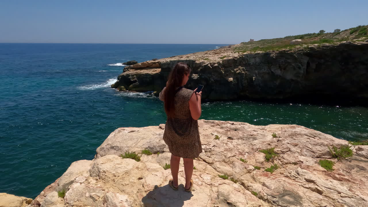 mujer de pie en el acantilado con un teléfono móvil en la costa oeste de malta, españa
