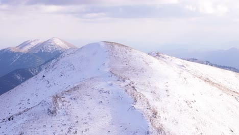 Schneebedeckter-Berggipfel-In-Leichter-Staubdecke-über-Felsigem-Gelände,-Abstieg-Aus-Der-Luft