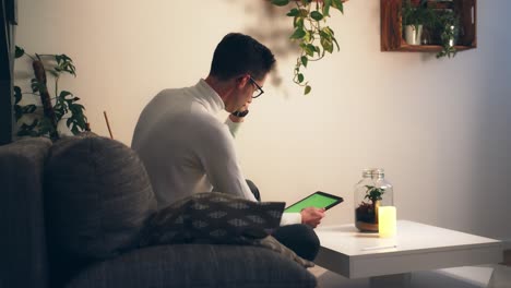 Plano-Medio-Completo-De-Un-Joven-Sentado-En-Un-Sofá-En-Casa-Usando-Una-Tableta-Digital-Electrónica-Con-Pantalla-Verde