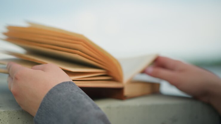 Closeup back view of an individual wearing grey clothing holding an open book with their right hand resting on the page while the wind softly flips through the pages, with blur background