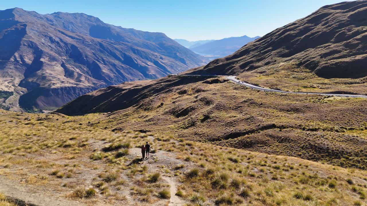 A couple walks through alpine scenery to enjoy views of Crown Range Road, the highest road in New Zealand. Mountains, winding roads, and valleys create a stunning natural scene