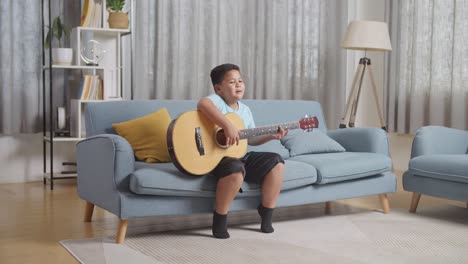 full body of talented asian kid enjoys singing and playing the guitar on sofa at home