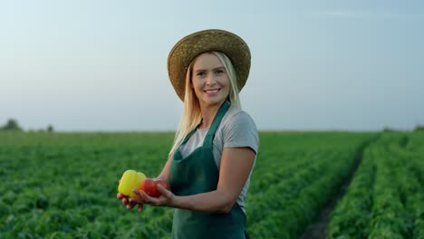 Portrait-Shot-Of-The-Beautiful-Young-Blond-Woman-In-A-Hat-And-Apron-Standing-In-The-Green-Field-And-Posing-To-The-Camera-With-Harvested-Vegetables-In-Hands-Like-Demonstrating-Them