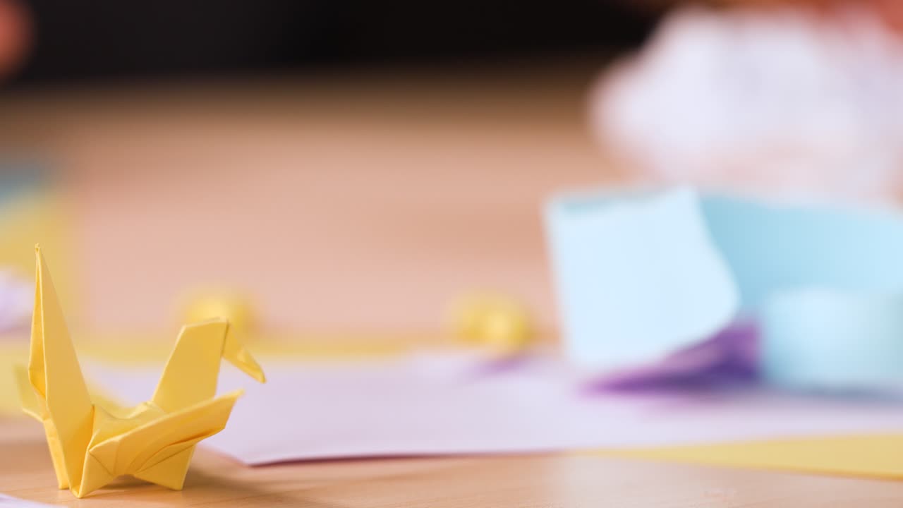 Una gru di carta gialla è appoggiata su una scrivania, mentre sullo sfondo delle mani piegano origami