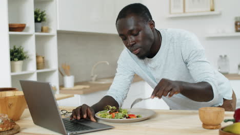 Young-Black-Man-Eating-Salad-and-Using-Laptop-in-Kitchen