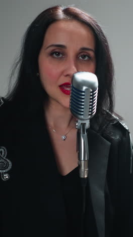 vocalista femenina enfocada en negro actuando con un micrófono vintage, guitarrista en el fondo, contra un telón de fondo de estudio blanco