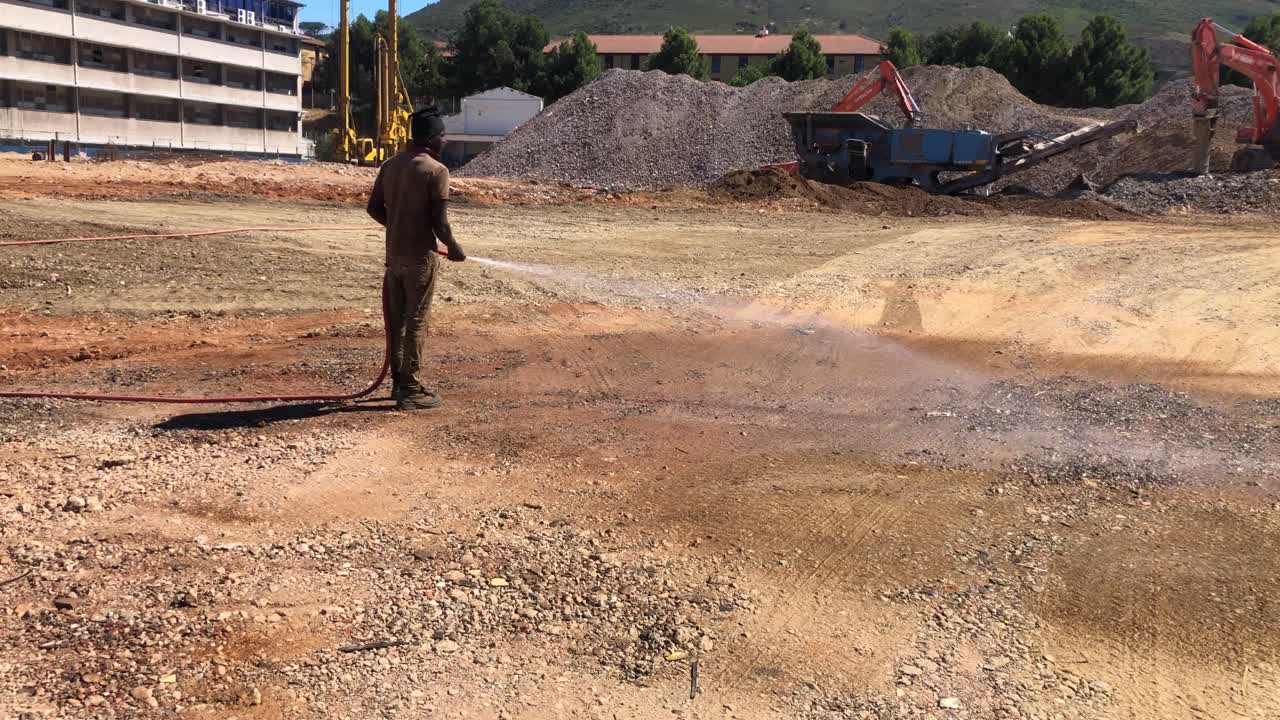 wide shot of worker watering the dry and dusty ground of a construction site in Cape Town, South Africa and preparing it for the use with heavy machinery like the Hitachi ZAXIS.