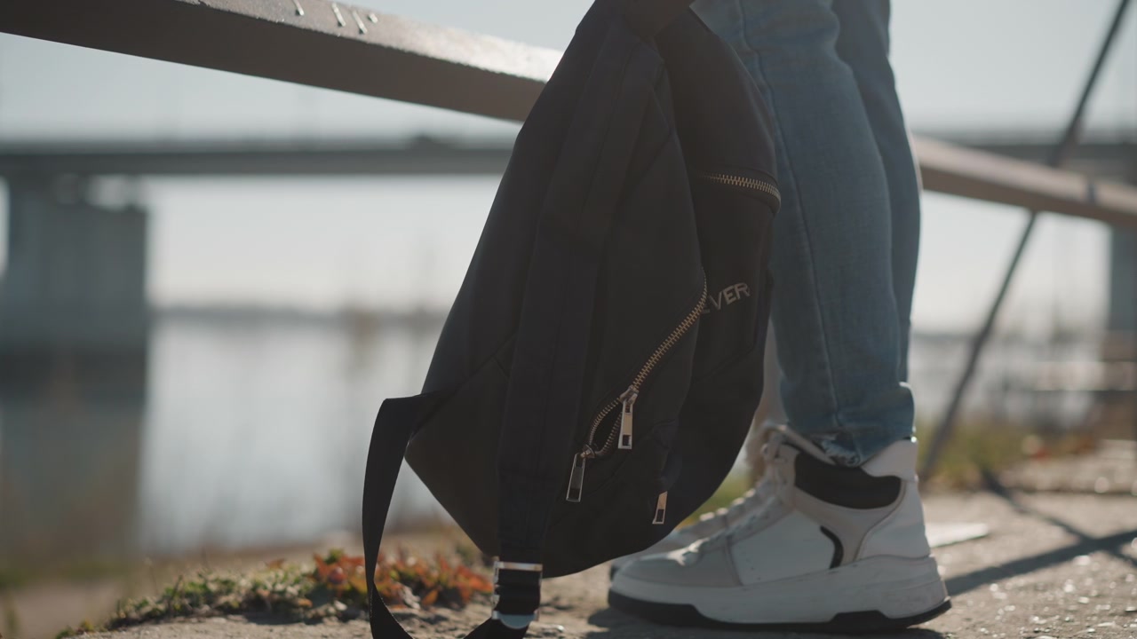 Donna in piedi vicino a una ringhiera metallica, con le gambe e uno zaino nero visibili, indossa jeans blu e scarpe da ginnastica bianche. Lo sfondo presenta un ponte sfocato sull'acqua in condizioni di luce diurna.