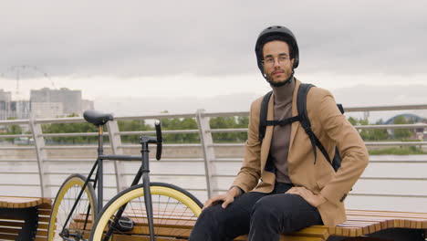 gutaussehender amerikaner in formeller kleidung mit helm und rucksack, der in die kamera schaut, während er neben seinem fahrrad auf einer bank auf der stadtbrücke sitzt